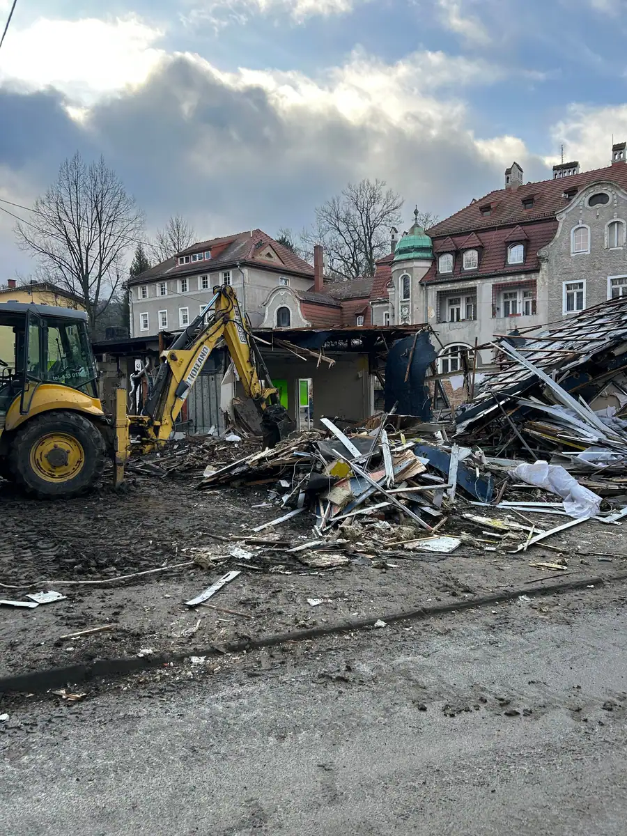 Koparko-ładowarka New Holland LB115B przy rozbiórce pawilonu, sterta desek i blachy